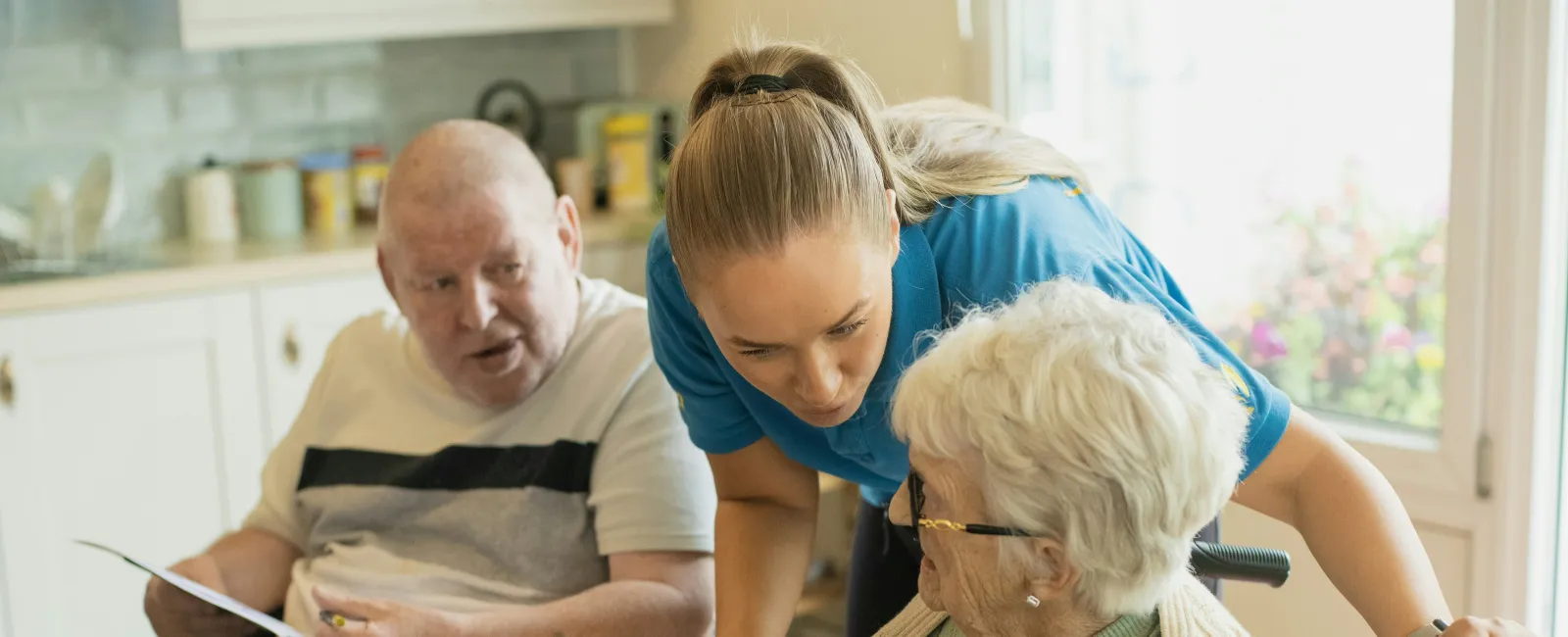 Caregiver assisting elderly woman in wheelchair with coloring activity while elderly man watches in bright room.