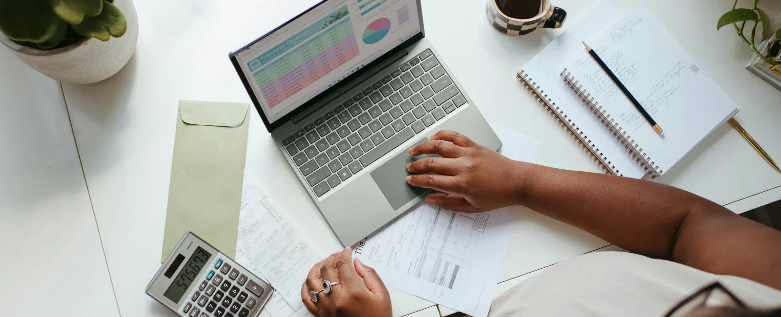 Person working on laptop with financial spreadsheets, calculator, documents, notebook, and coffee on a white desk.