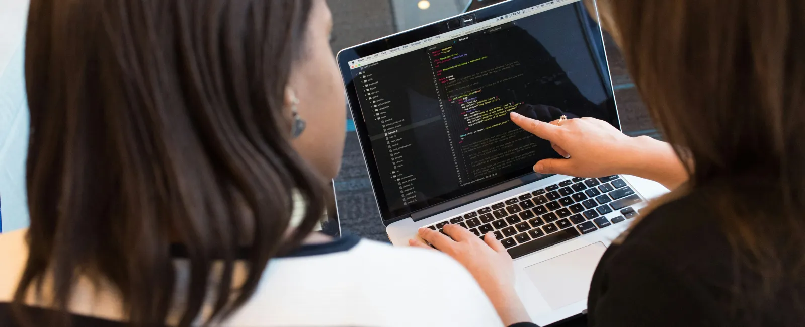 Two women collaborating on coding by reviewing programming code on a laptop screen in a modern workspace.