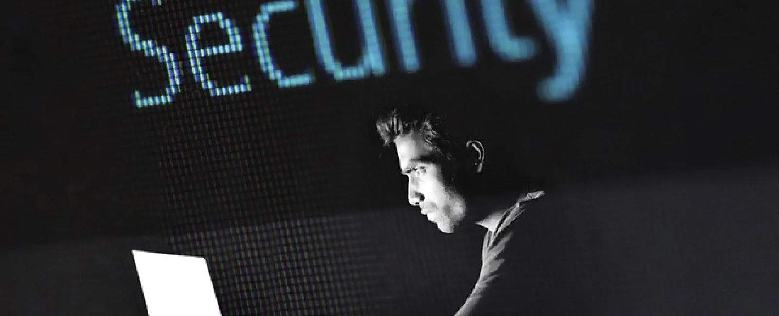 Man working on laptop with the word Security projected in large blue letters behind him in dark setting