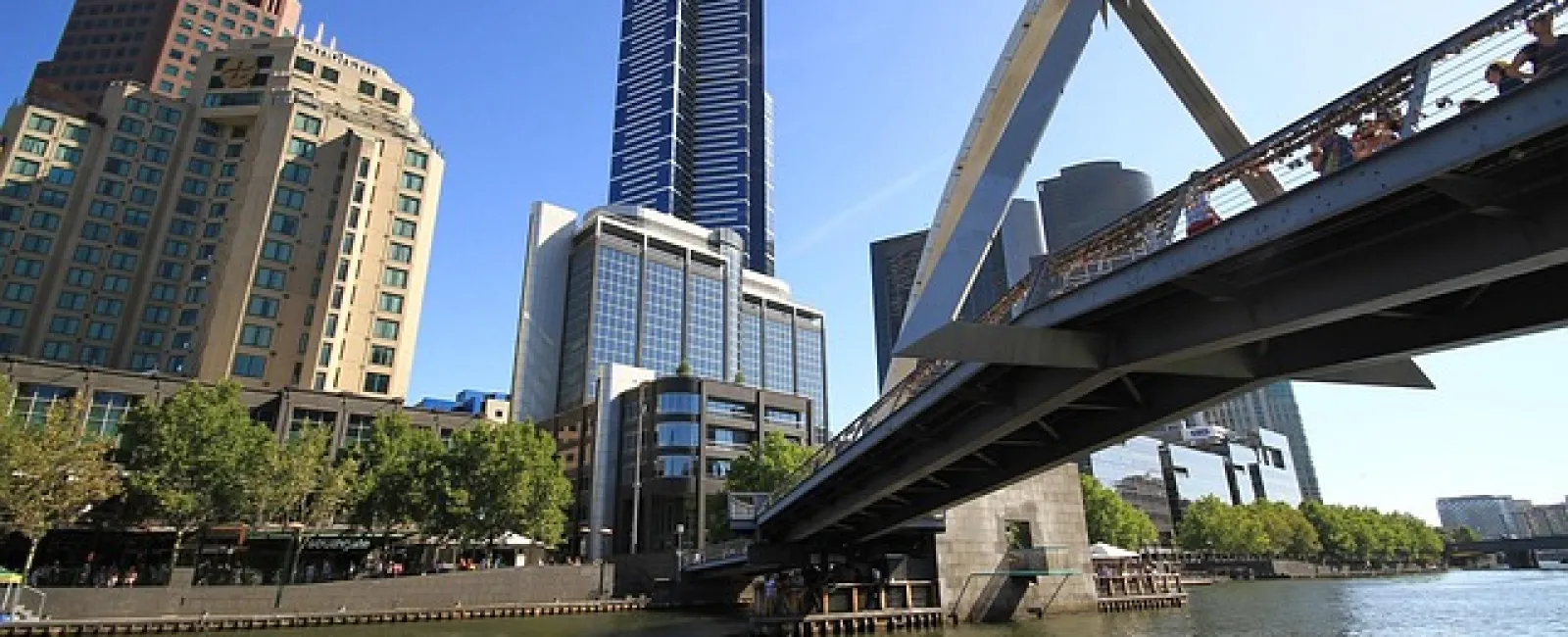 View of a modern city skyline with a tall skyscraper, riverside buildings, and a pedestrian bridge over water.
