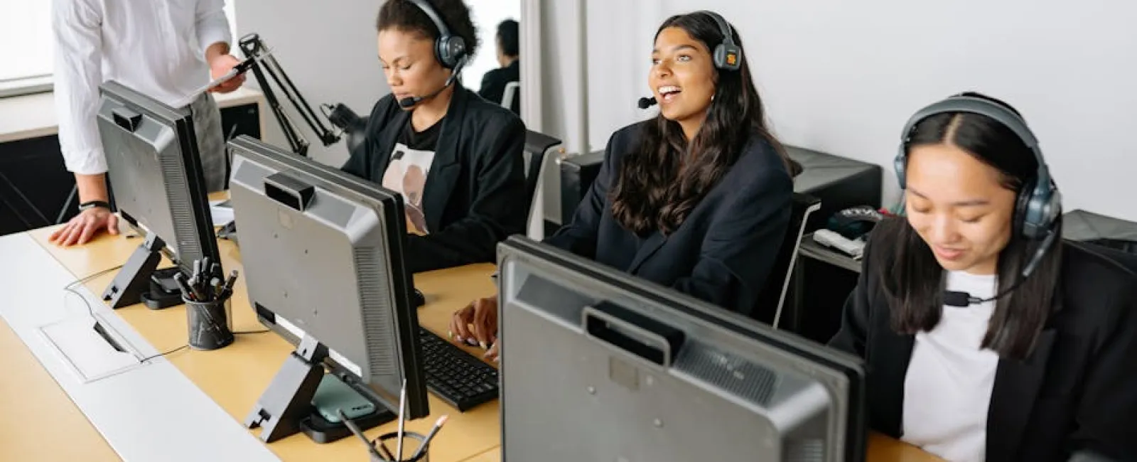 Diverse call center team with headsets working on computers and a male supervisor assisting staff in a modern office.