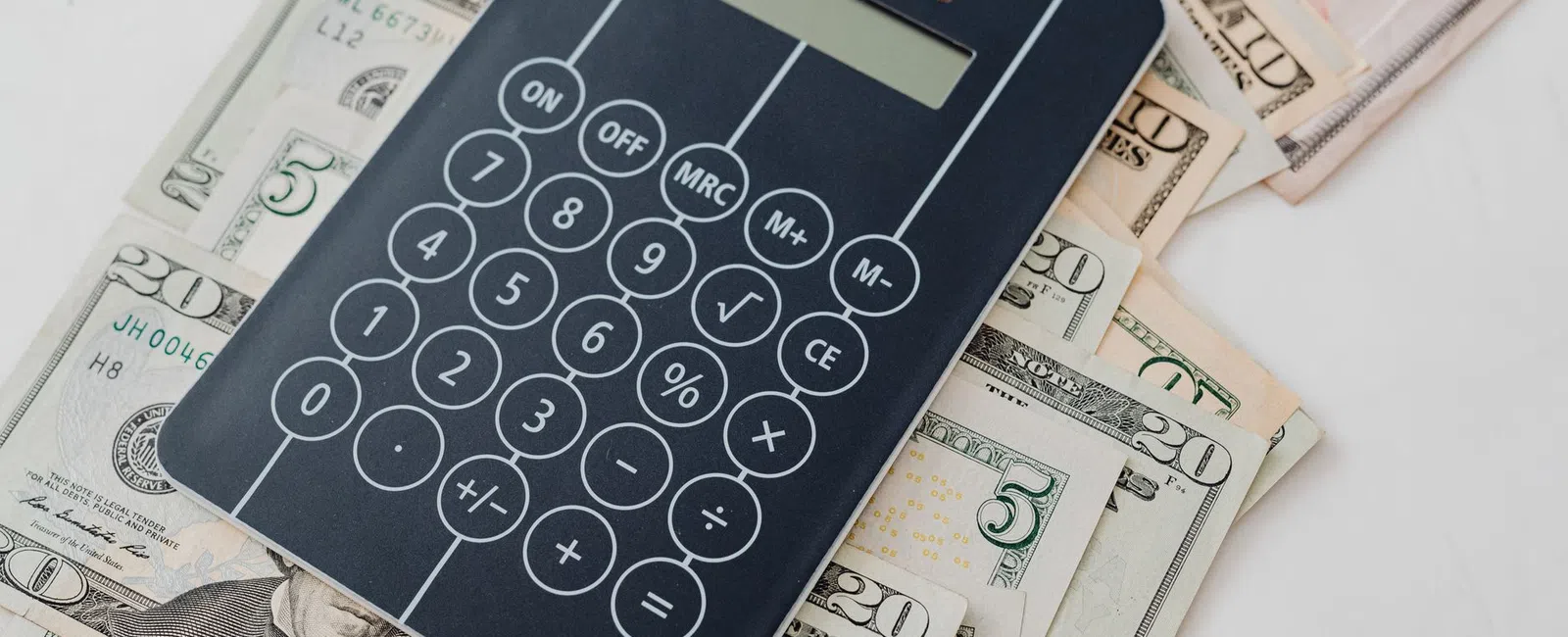 Black calculator on stacked US dollar bills of various denominations on a white surface.