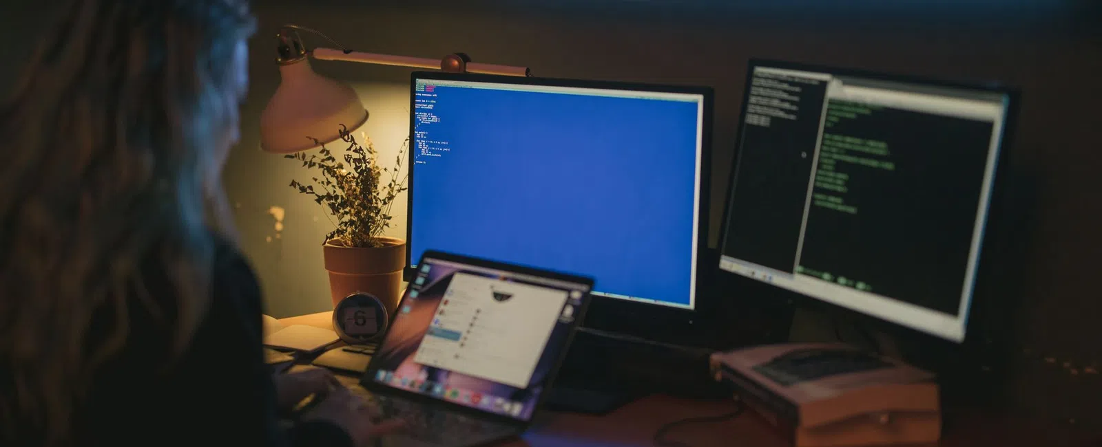 Person working on a laptop with two monitors displaying code in a dimly lit workspace with plants and a desk lamp.