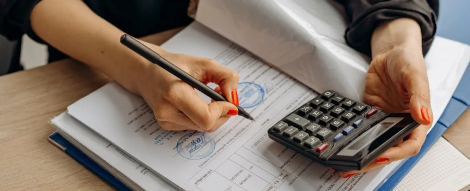 Person calculating finances with a calculator and pen while filling out official documents on a desk.