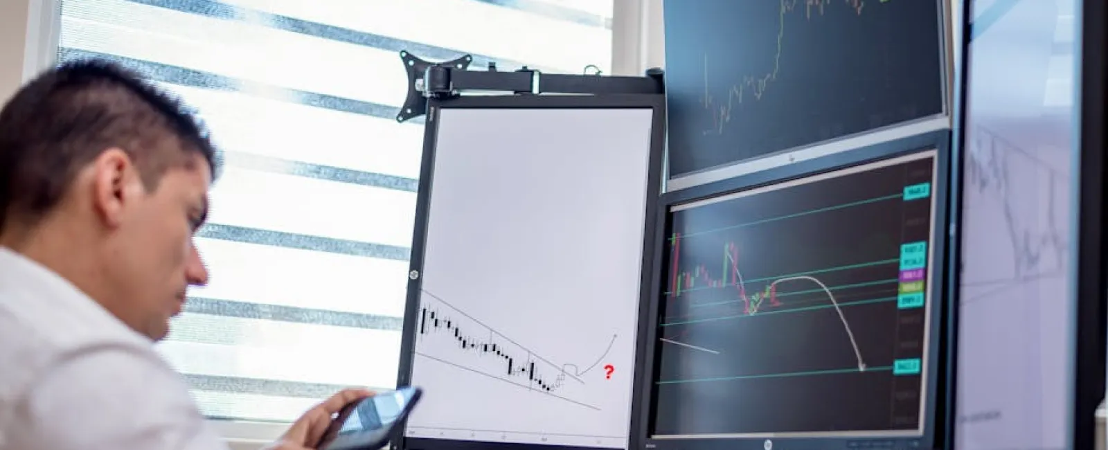 Man analyzing stock charts on multiple monitors and smartphone in a bright office setting
