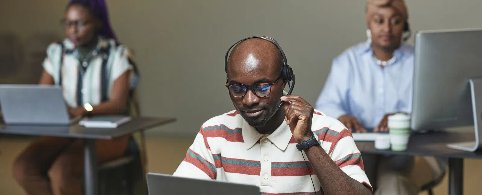 Three diverse professionals working on computers in an office, man in headset in foreground focused on laptop.