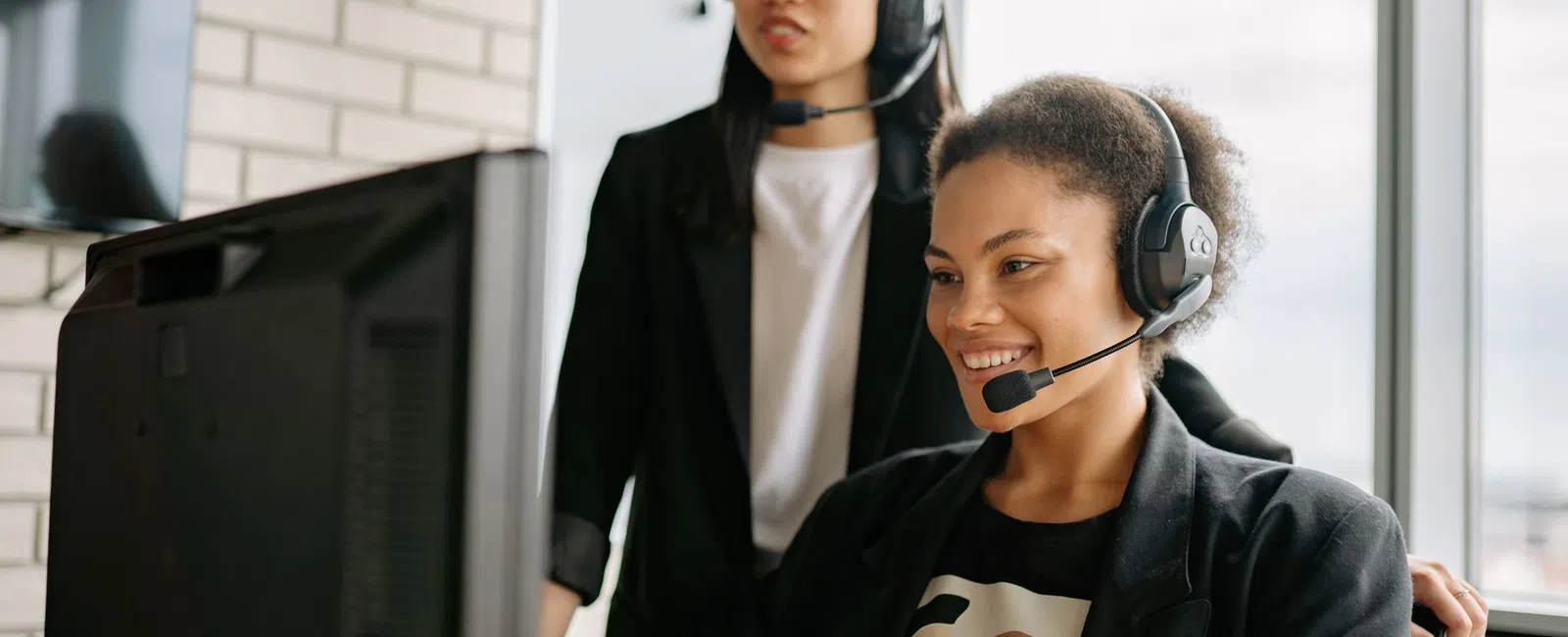 Two female customer service agents wearing headsets working together at a computer in an office.