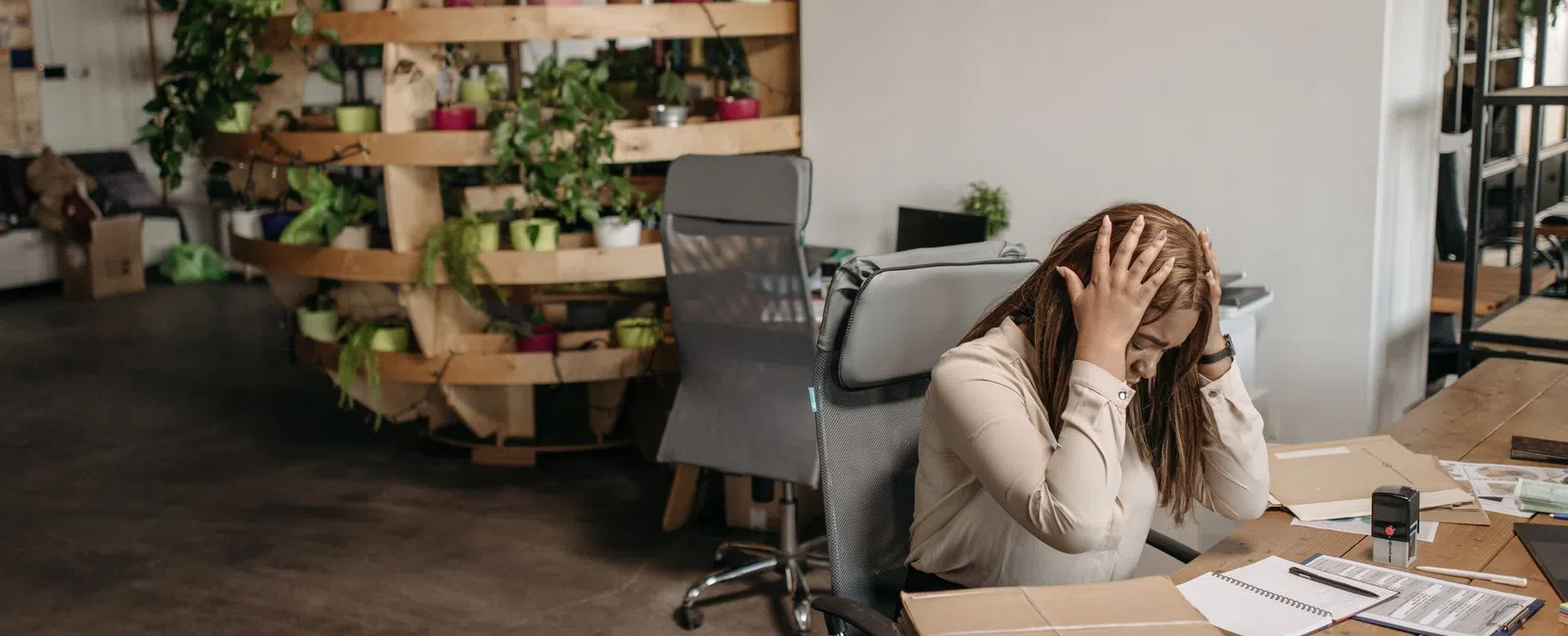 Stressed woman sitting at desk holding head with paperwork and files in a modern office with plants.