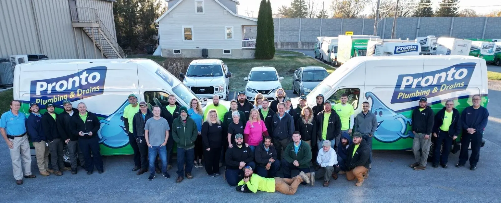Large group of Pronto Plumbing & Drains employees posing in front of company vans in a parking lot