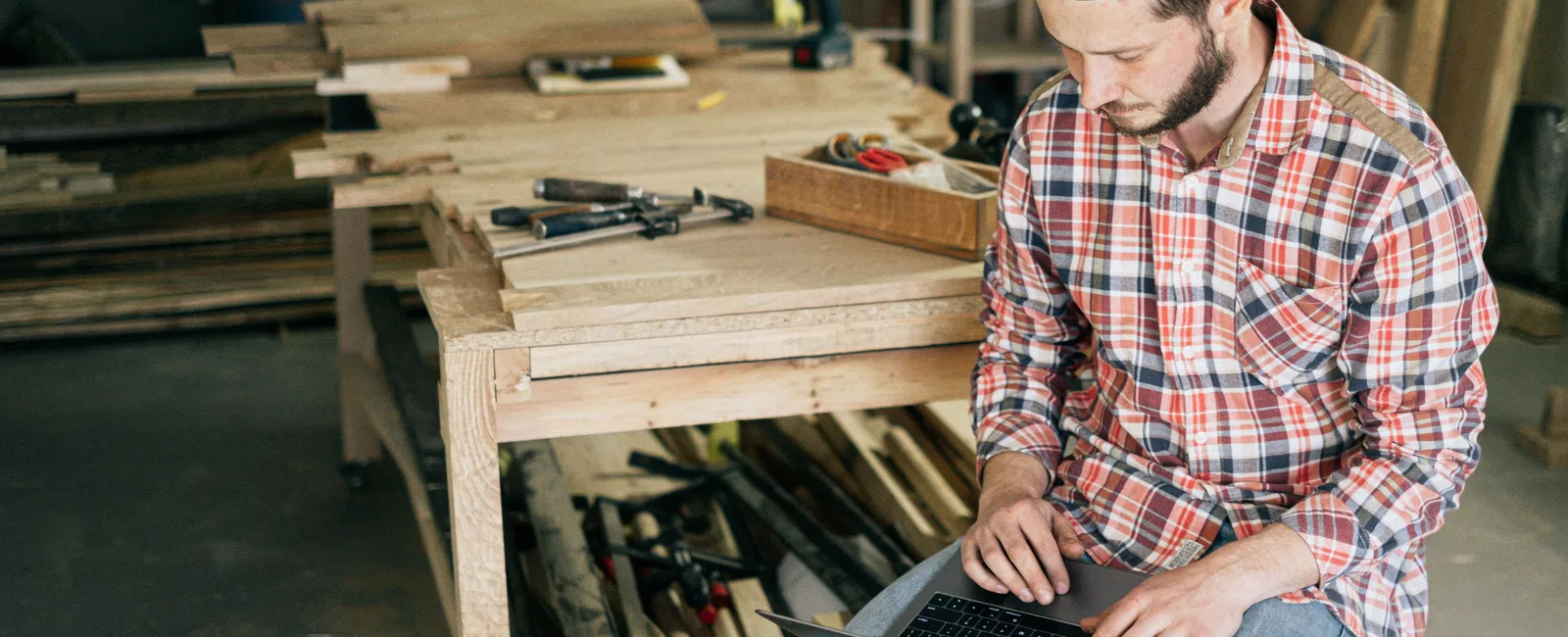 Carpenter in plaid shirt working on laptop in a woodworking shop surrounded by tools and wood materials