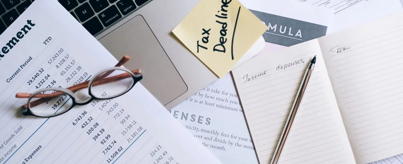Workspace with tax documents, glasses, laptop, sticky note reading Tax Deadline, and notebook with pen