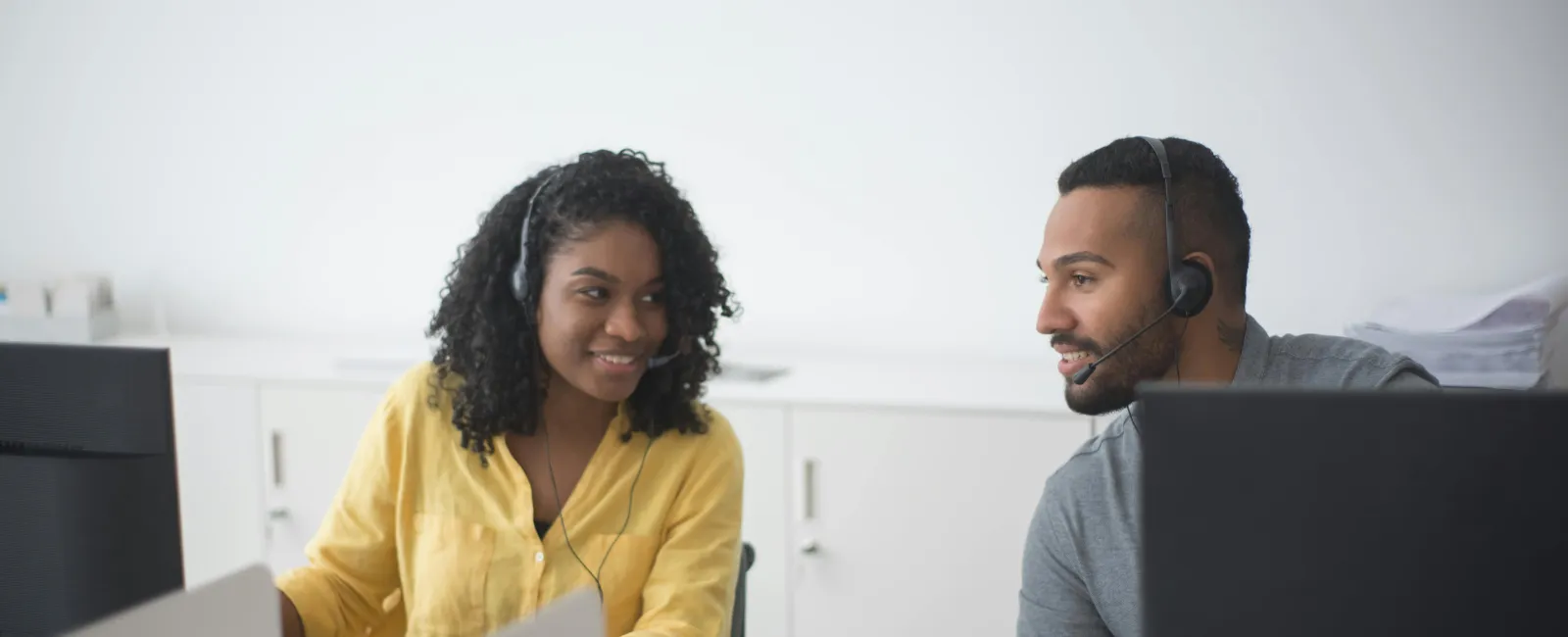 Two customer service representatives wearing headsets assisting clients at computers in an office setting.