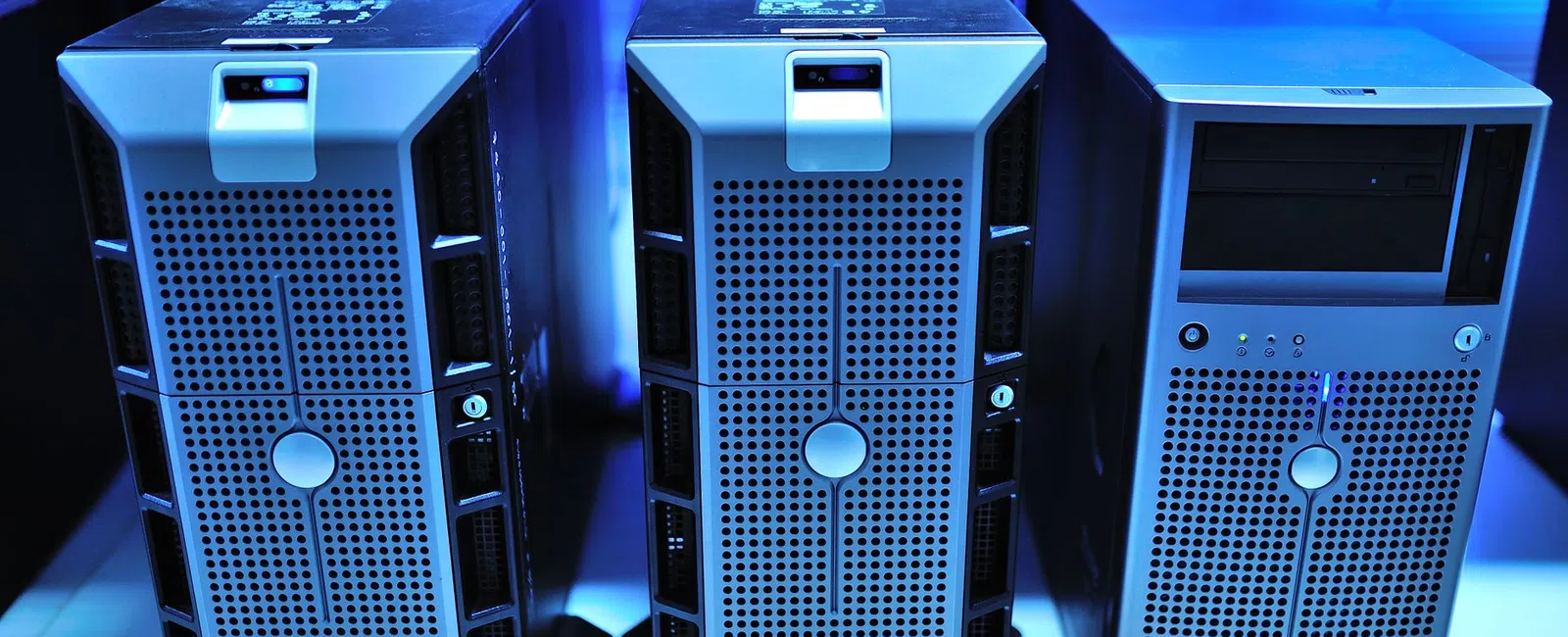 Three silver computer servers with perforated front panels on a white shelf under blue lighting.