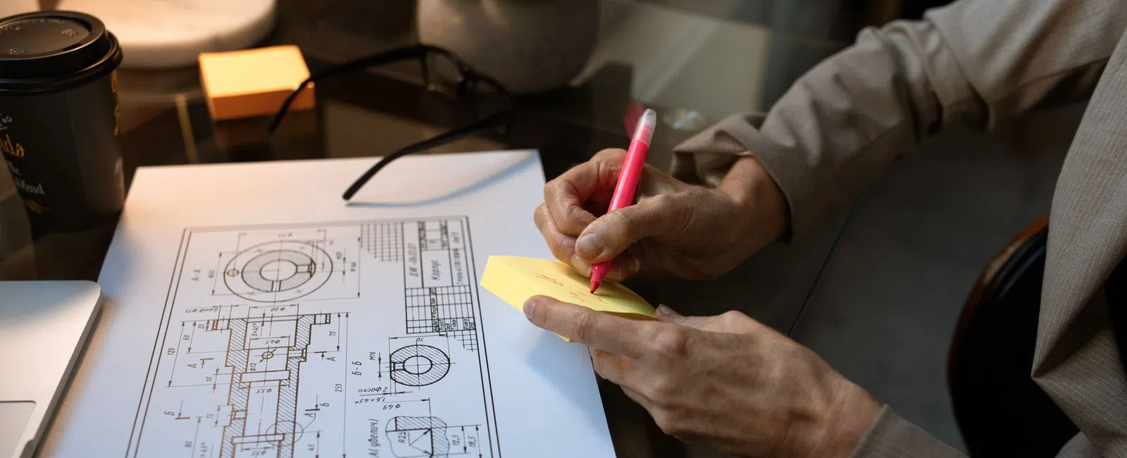Person writing on yellow sticky notes next to engineering blueprint and workspace items on glass desk