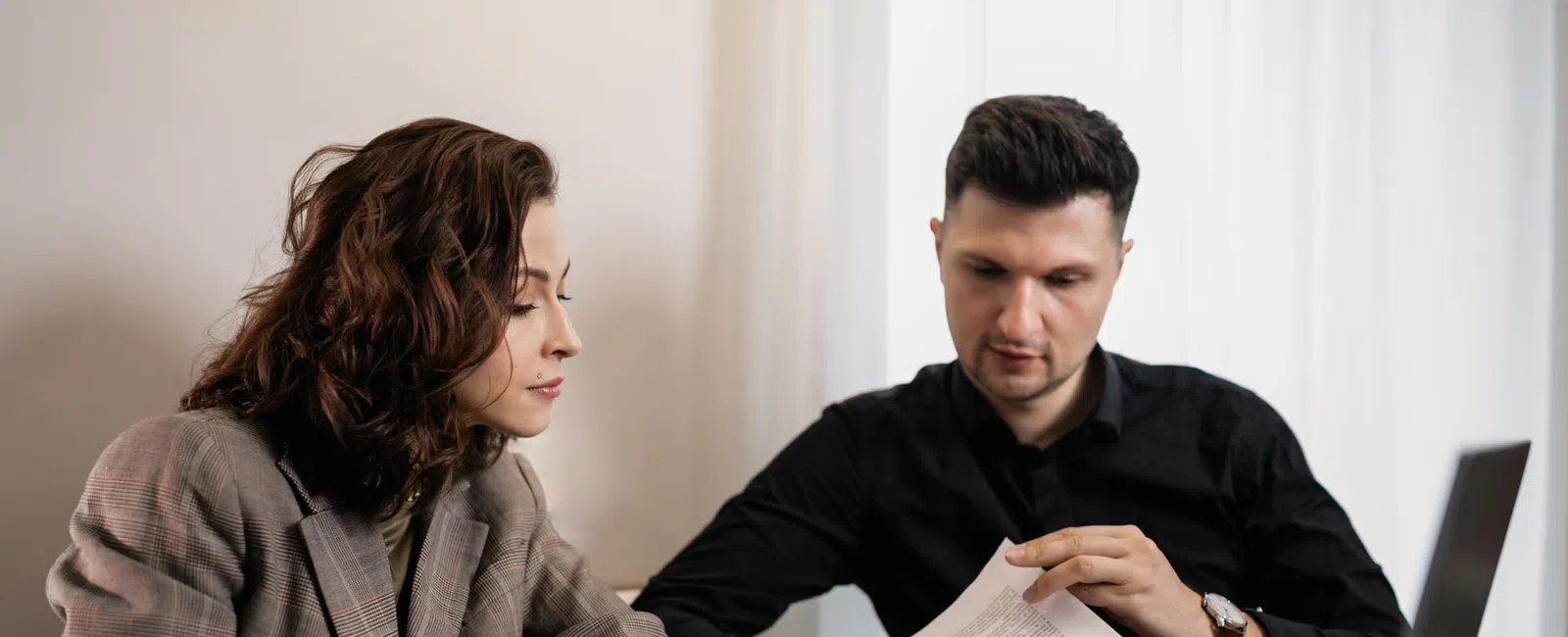 Two professionals discussing paperwork at a wooden table with a laptop in a modern office setting.
