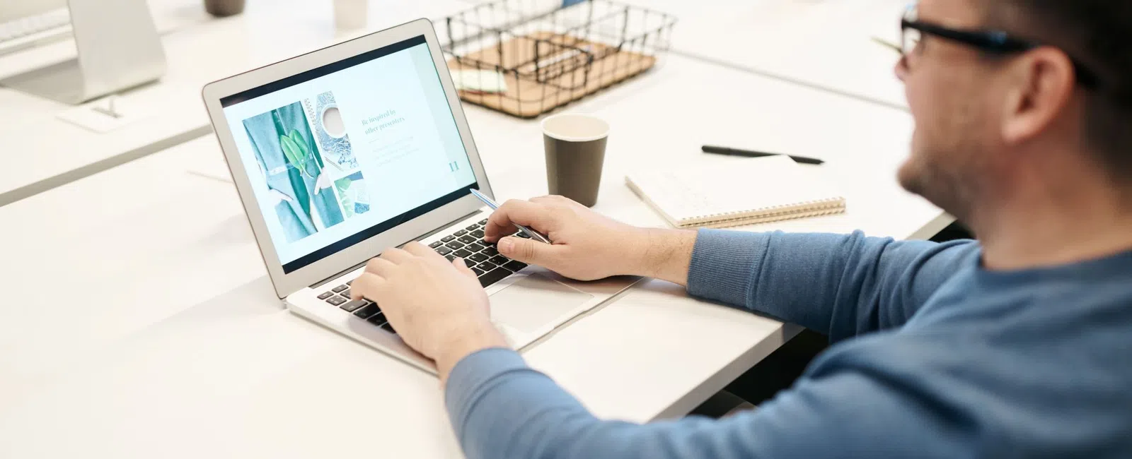 Man in blue sweater working on laptop in a bright office with coffee cup and notepad on desk.