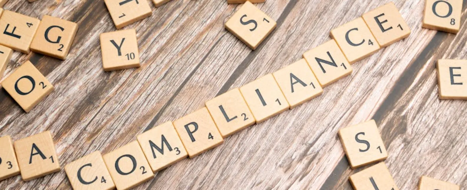 Wooden Scrabble tiles spelling out the word compliance on a wooden surface with scattered letter tiles around.