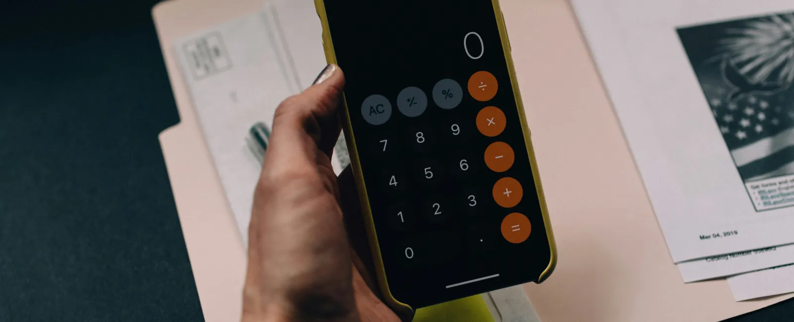 Hand holding smartphone displaying calculator app over IT budget documents.