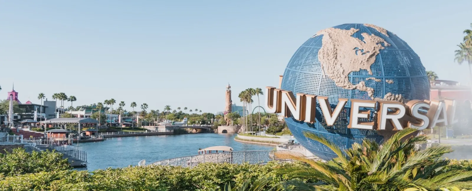 Large Universal Studios globe sculpture near lush greenery with water and buildings in the background under clear sky.