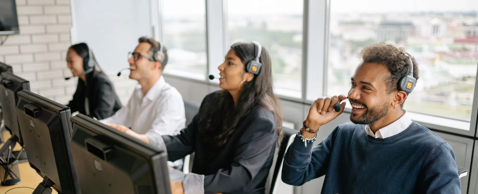 Diverse call center team wearing headsets working at computers in bright office with large windows.