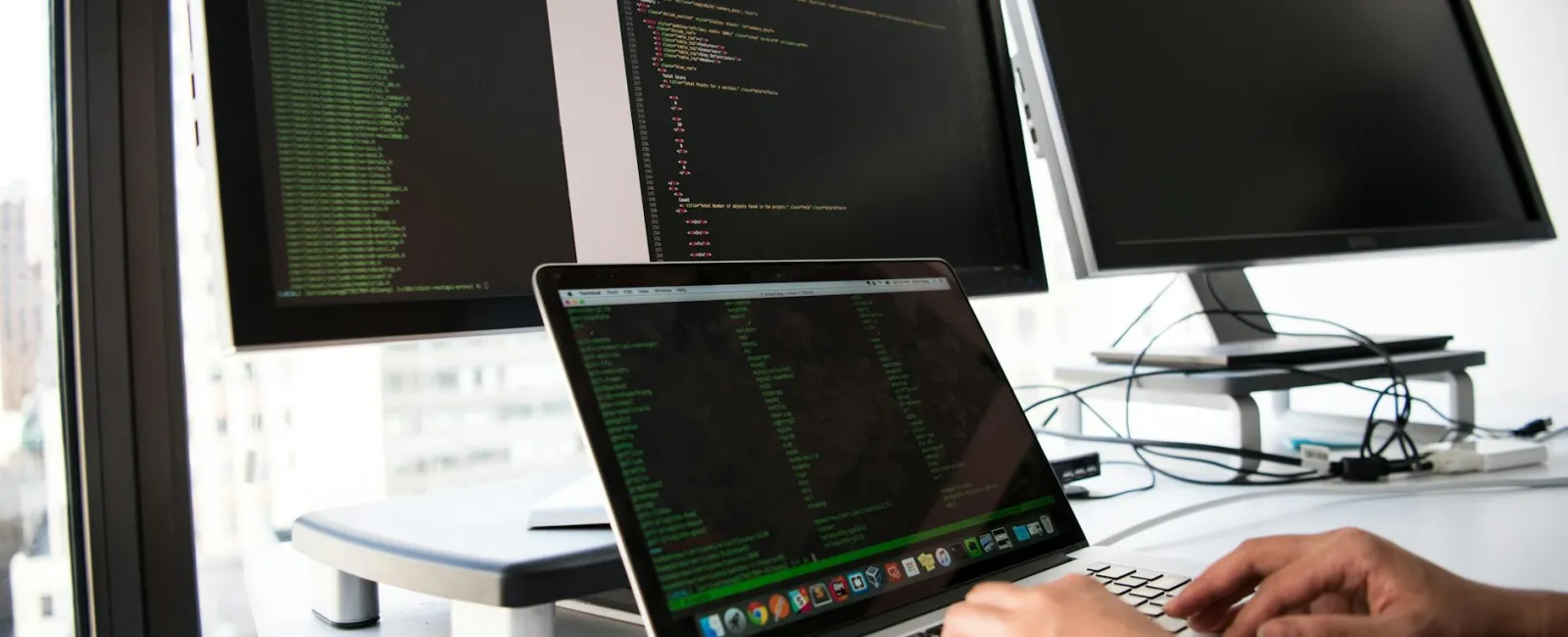Person typing code on laptop with two large monitors displaying programming scripts in a bright office space.