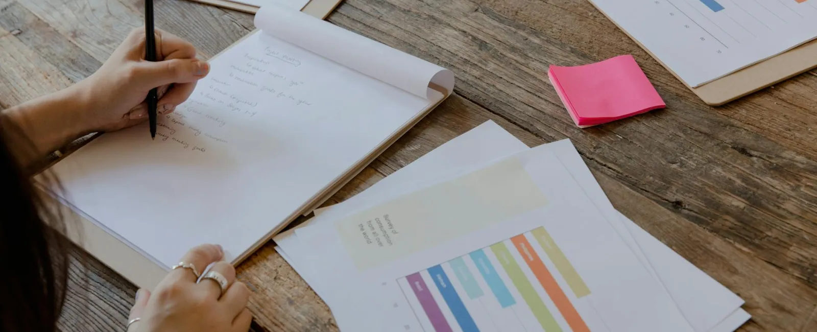 Person writing notes on a notepad with project charts and colorful bar graphs on wooden table
