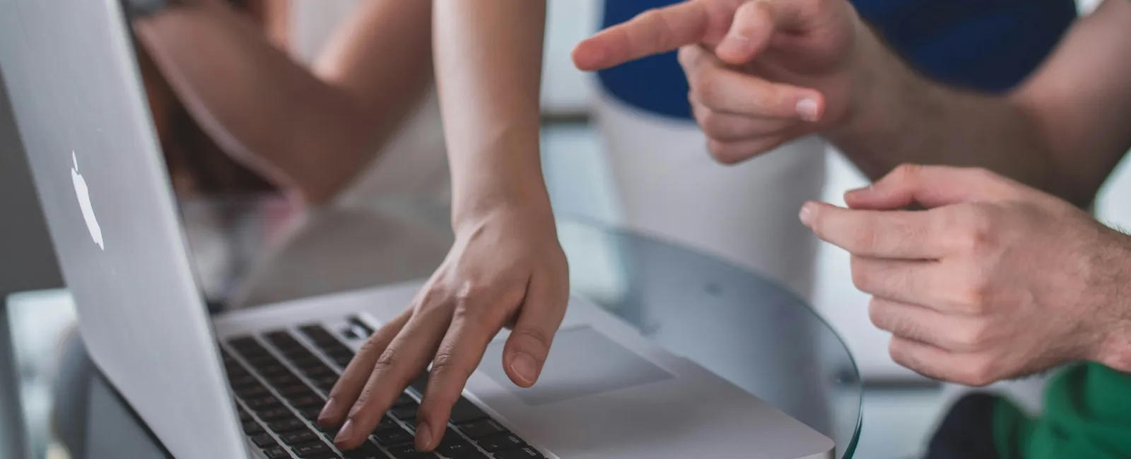 People collaborating over a laptop on a glass table, with hands pointing and typing on the keyboard.