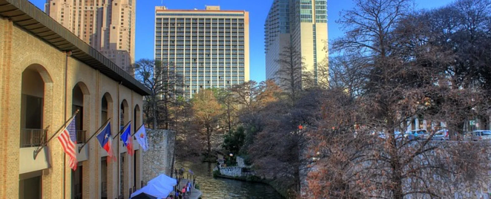 San Antonio Riverwalk with flags, tents, and city skyscrapers under a clear blue sky