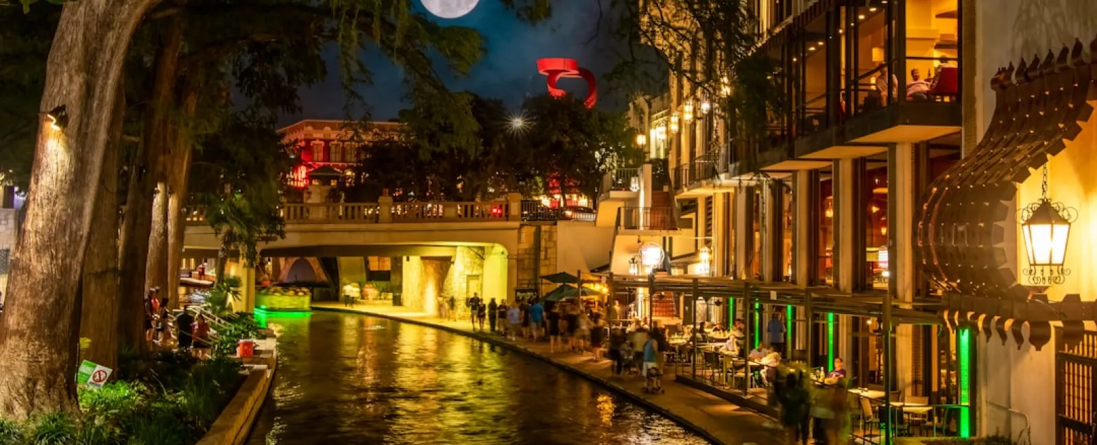 Night view of a lively riverwalk with illuminated restaurants, a full moon, and reflections in the water