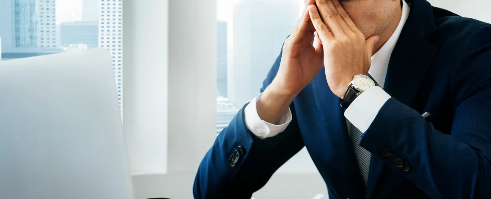 Stressed businessman in office rubbing eyes with glasses, laptop, crumpled paper on desk, city view background - the real cost of IT downtime