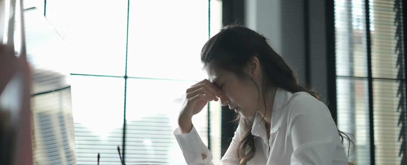 Stressed woman in white shirt sitting at desk with computer, holding forehead in office with large windows.