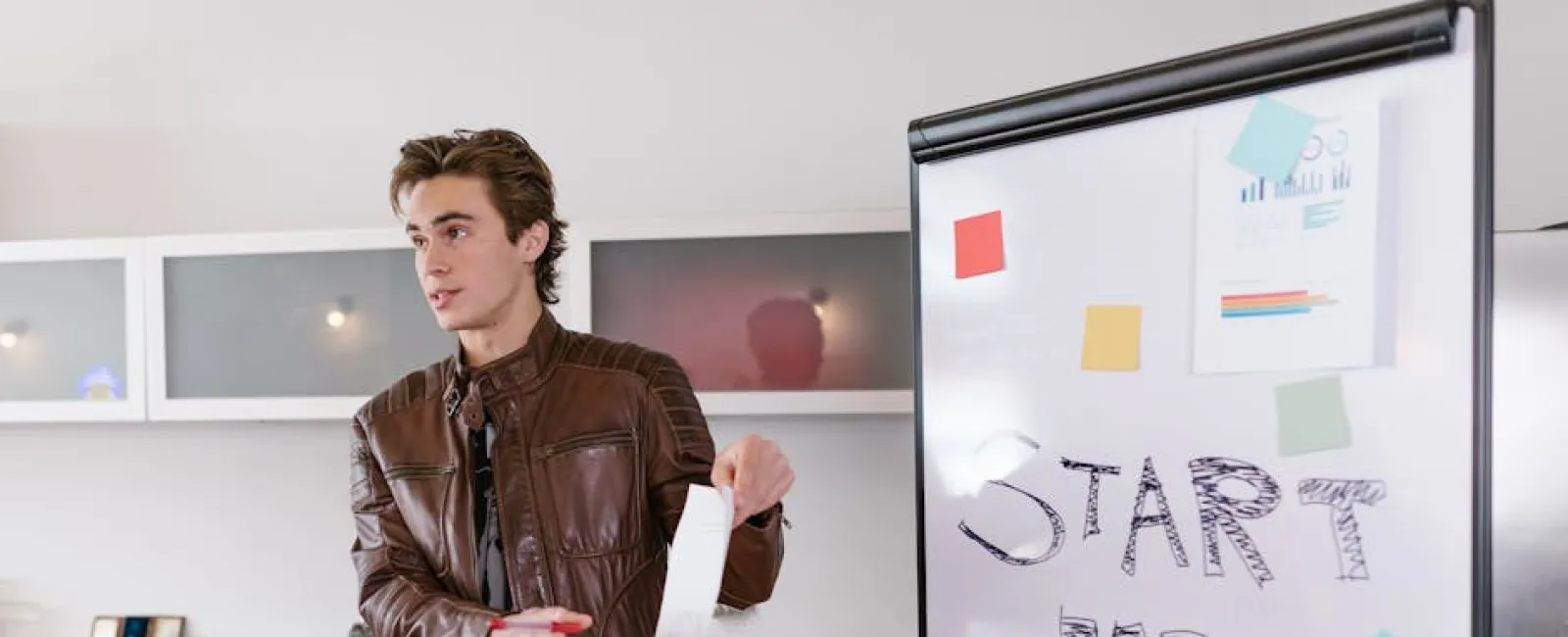 Young man in brown leather jacket presenting startup ideas on whiteboard with notes and charts in office.