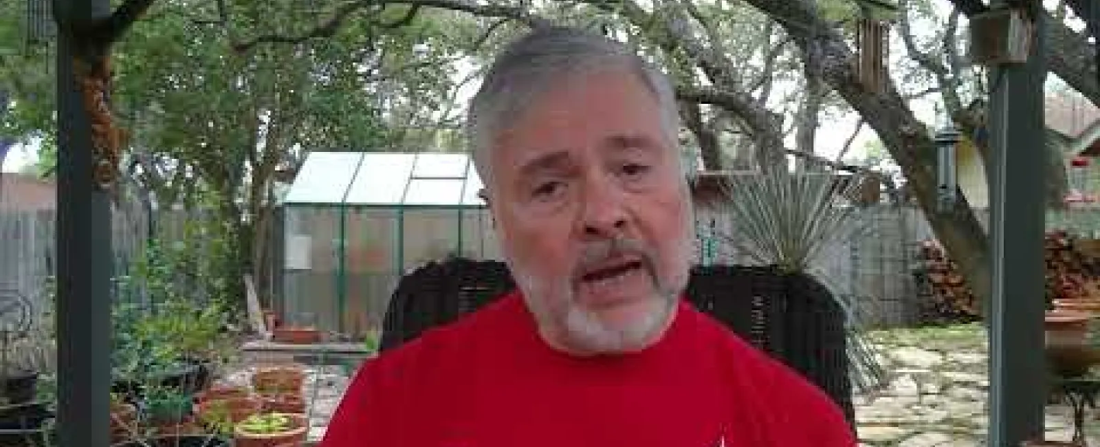 Man with gray beard wearing a red TechSage Solutions shirt sitting outdoors near plants under a wooden pergola.