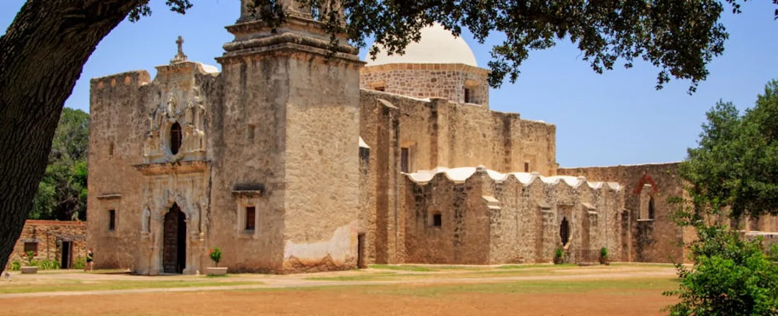 Historic stone mission building with dome and arched entrance under a large tree on a sunny day.