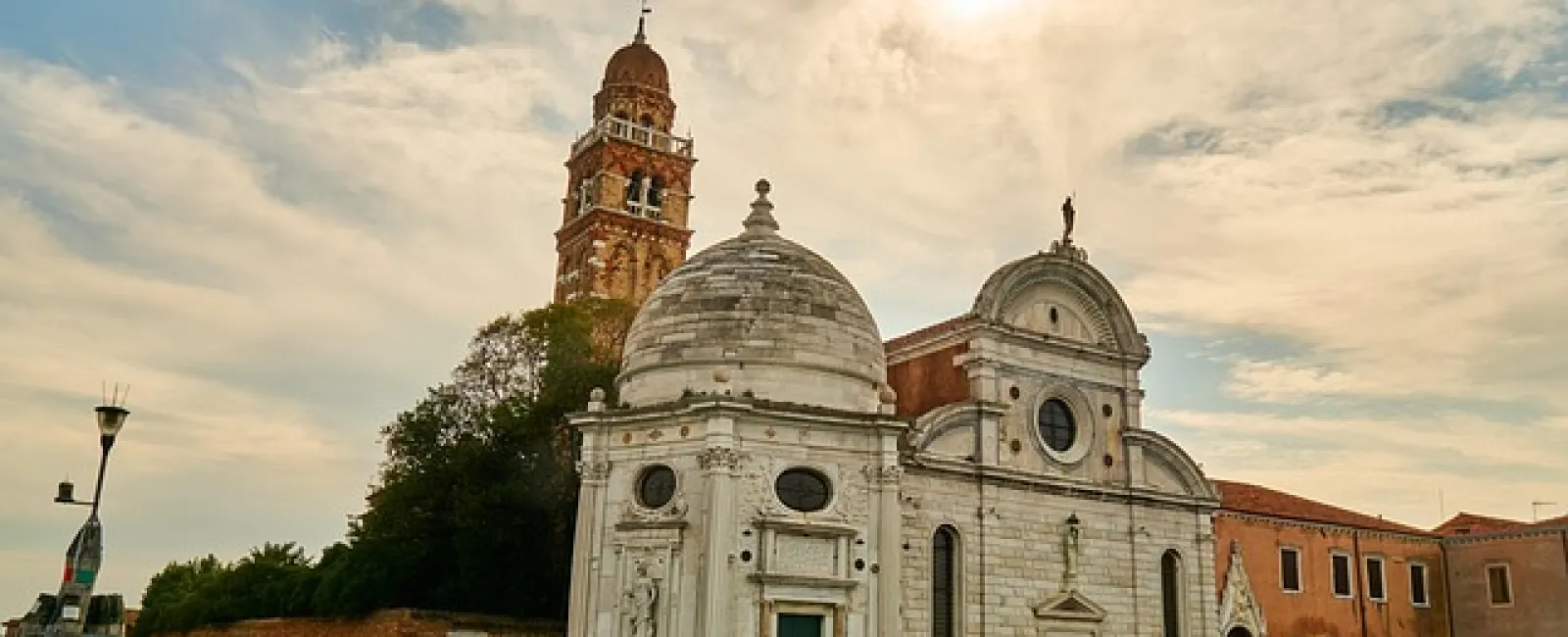 Historic white stone church with dome and bell tower beside canal under a cloudy sky at sunset.