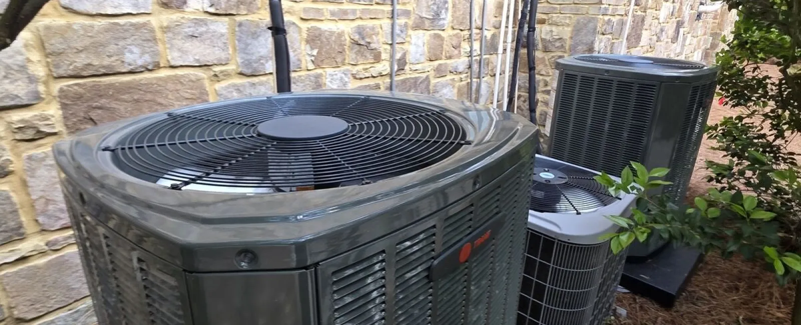 Outdoor air conditioning units installed next to a stone brick exterior wall surrounded by landscaping and trees.