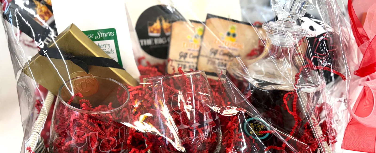 Gift basket wrapped in clear plastic with red shredded paper, glasses, snacks, and a bottle with red ribbon