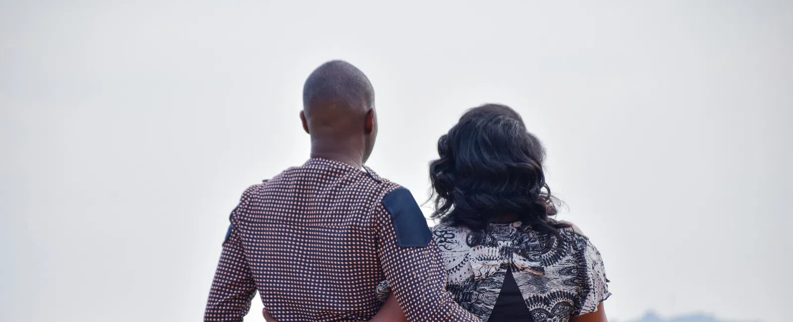 Couple embracing by a waterfront with calm water and distant shore under clear sky.