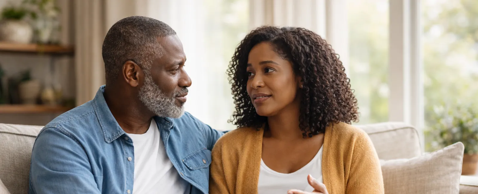 Mature man and young woman sitting on couch engaged in a heartfelt conversation at home.