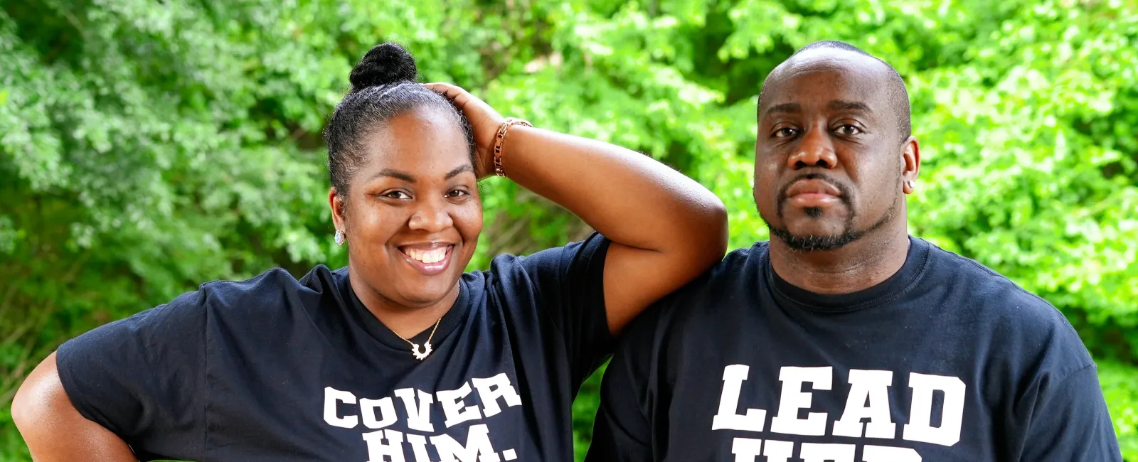 A happy couple outdoors wearing matching black shirts with messages Cover Him and Lead Her in white text.