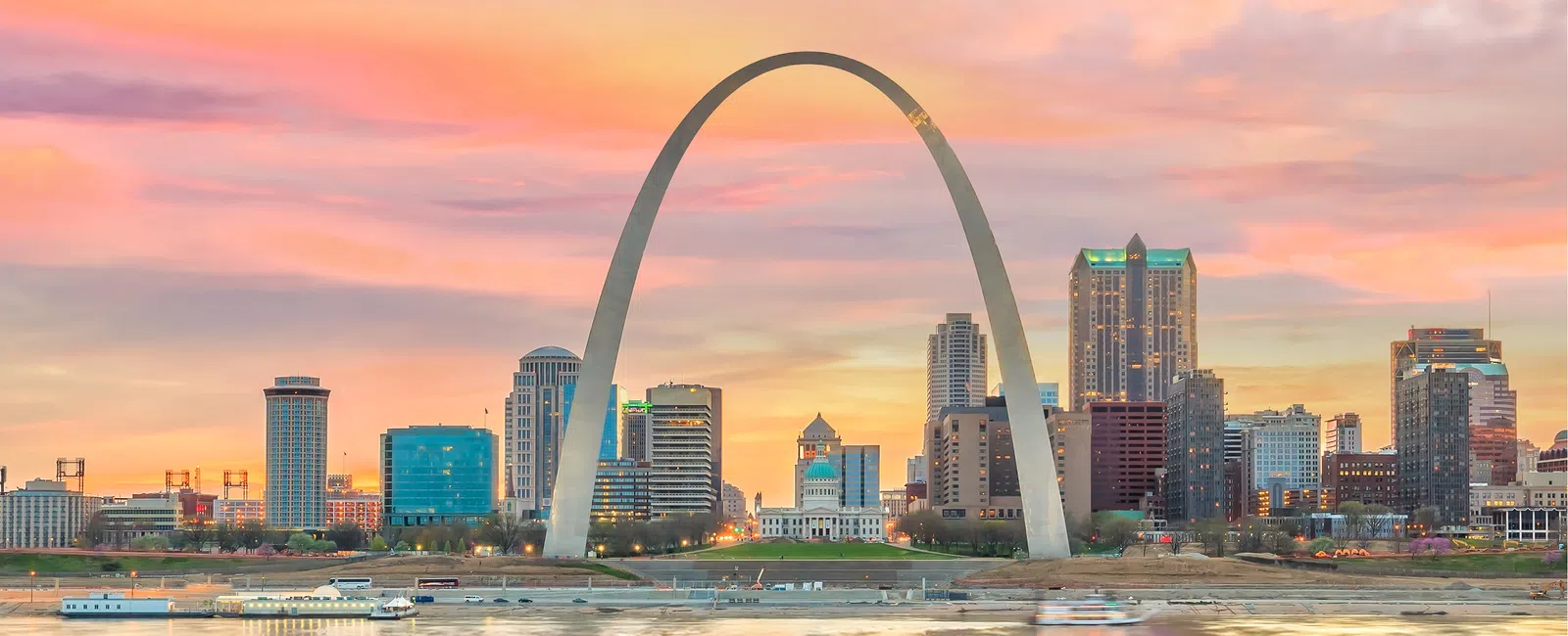 St. Louis Gateway Arch at sunset with city skyline and river reflecting colorful sky hues.