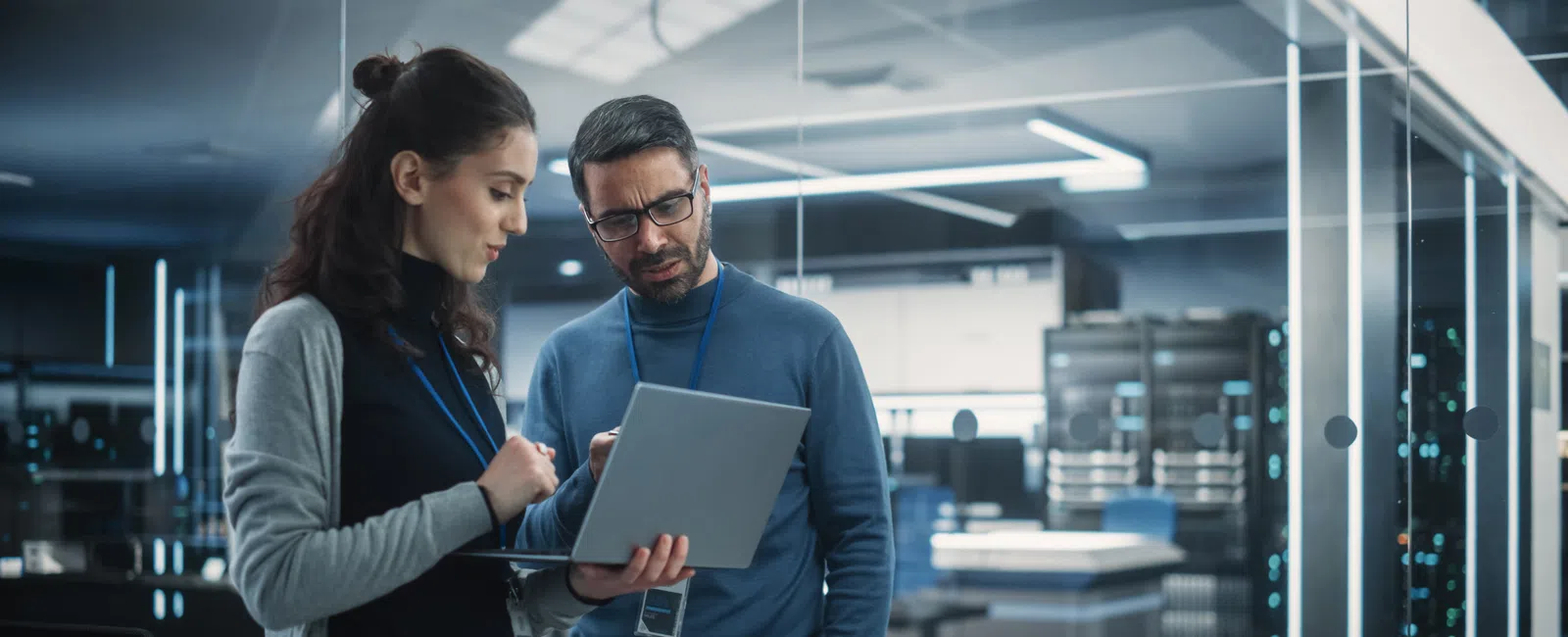 Two IT professionals discuss data on a laptop inside a modern, high-tech server room with visible racks and equipment.