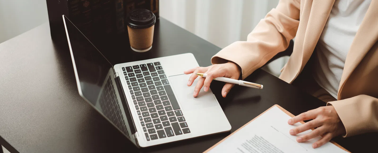 Person in beige blazer working on laptop with clipboard and coffee on desk in office setting.
