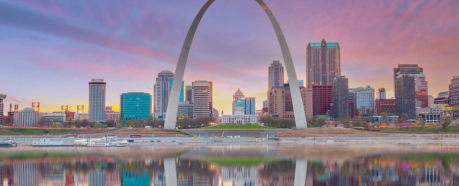 St. Louis Gateway Arch and downtown skyline reflected in the Mississippi River during a colorful sunset.
