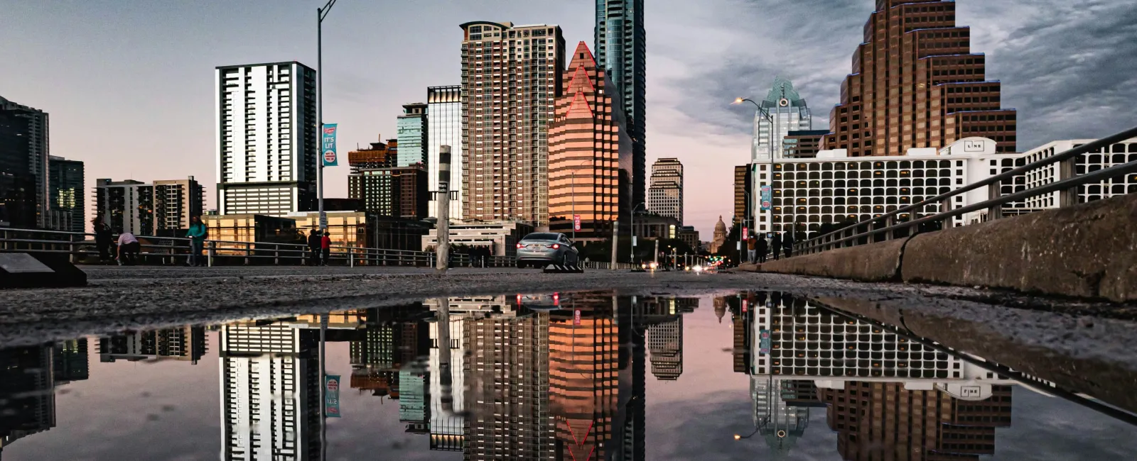 City skyline with tall buildings reflected in a large puddle on a street during dusk with cloudy sky.