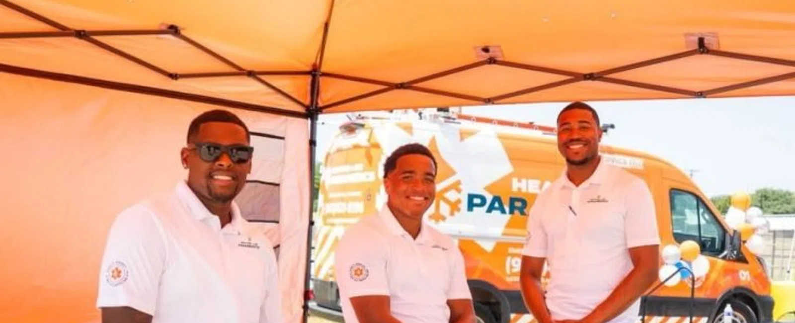 Three men in white shirts and blue shorts under a tent with a Heating Air Paramedics van outside on a sunny day