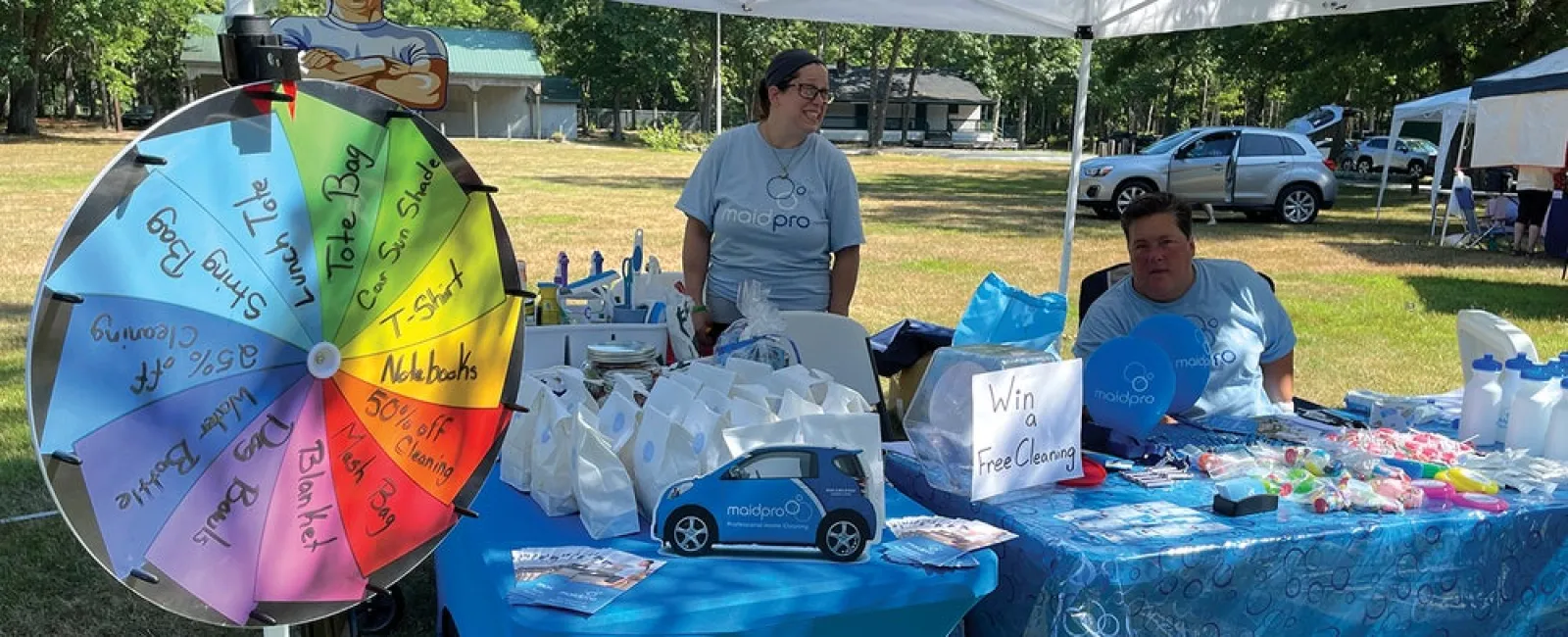 Event booth with prize wheel, promotional items, and two staff under white canopy at outdoor fair