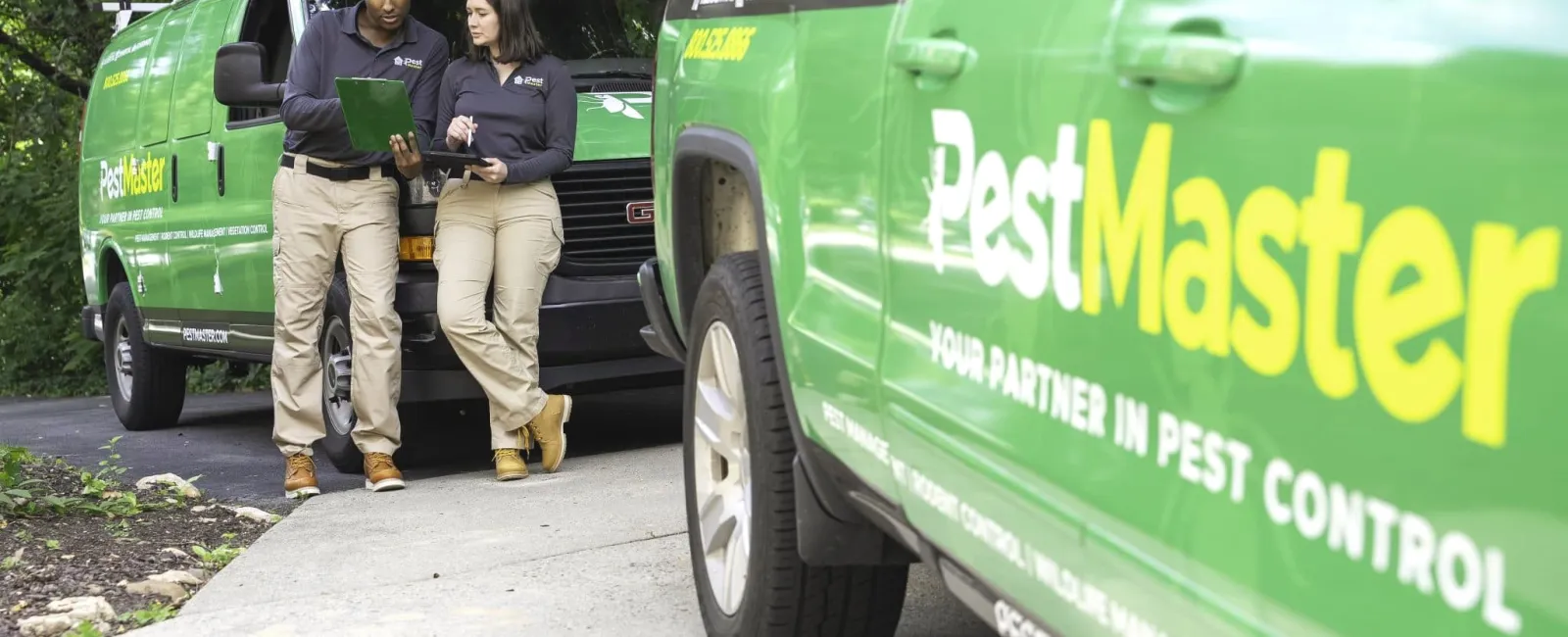 Pest control professionals reviewing documents beside green Pest Master service vehicles in outdoor setting.