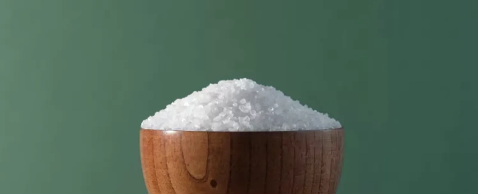 Coarse sea salt crystals in a wooden bowl against a green background on wooden surface